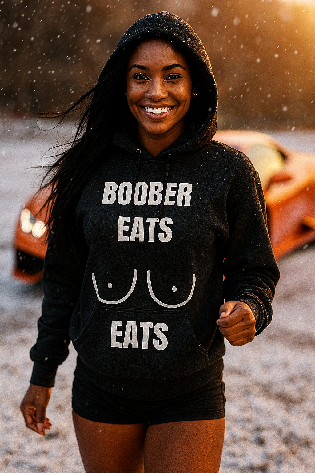 Multi-ethnic athletic woman (18–22) with dark skin, long black hair, and brown eyes, smiling waist-up while holding the drawstrings of her black Gildan 18500 hoodie that features the white “BOOBER EATS” graphic, standing in light snowfall during golden hour with an orange sports car blurred in the background.