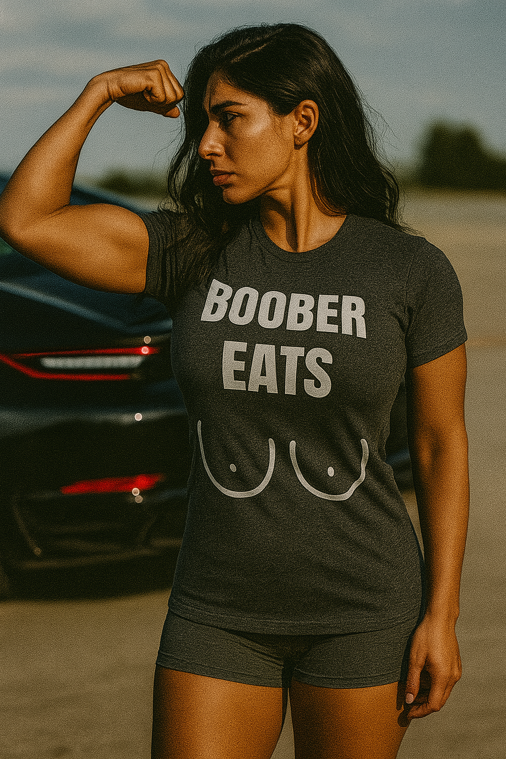 Strong Middle Eastern woman with wavy black hair flexing her bicep beside a luxury sports car while wearing a tailored-fit dark grey “Boober Eats” t-shirt and shorts; bold fitness apparel for traveling professionals like flight attendants and pilots.

