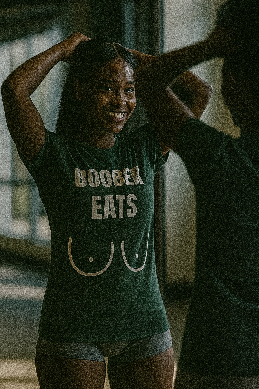 Athletic African-American woman with deep skin and straight long black hair smiling while tying her hair in front of a gym mirror, wearing a tailored-fit forest green “Boober Eats” t-shirt and shorts; bold travel fitness apparel for flight attendants, nurses, and pilots.

