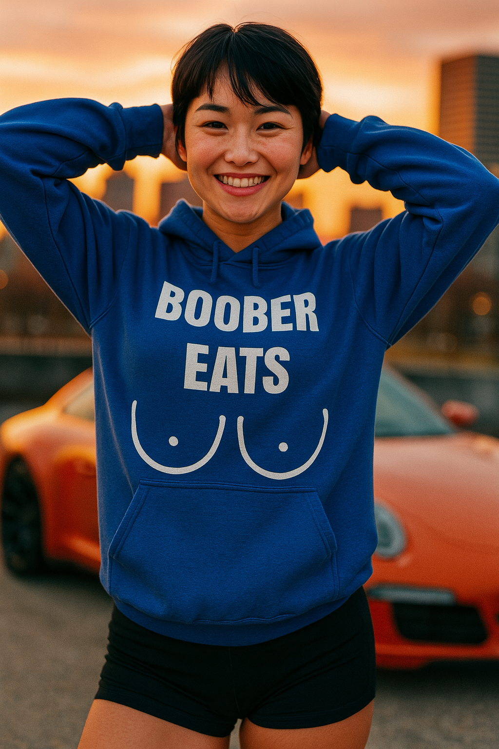 Athletic Asian woman (18–22) with fair skin, short black hair, and brown eyes, smiling waist-up with her hands resting on her head. She wears a tailored-fit indigo blue Gildan 18500 hoodie featuring the white “BOOBER EATS” graphic, paired with black athletic shorts. The dramatic sunset light casts warm tones over a blurred urban cityscape backdrop, giving the scene a cinematic, dynamic feel.

