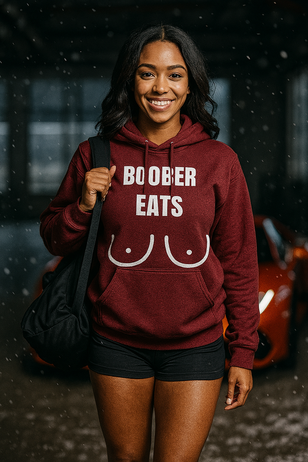 Light-skinned African American woman (18–22) with wavy black hair, brown eyes, and a bright smile, standing waist-up in a snowy gym interior while carrying a black duffel bag over her shoulder. She wears a maroon Gildan 18500 hoodie with the white “BOOBER EATS” graphic and black athletic shorts.

