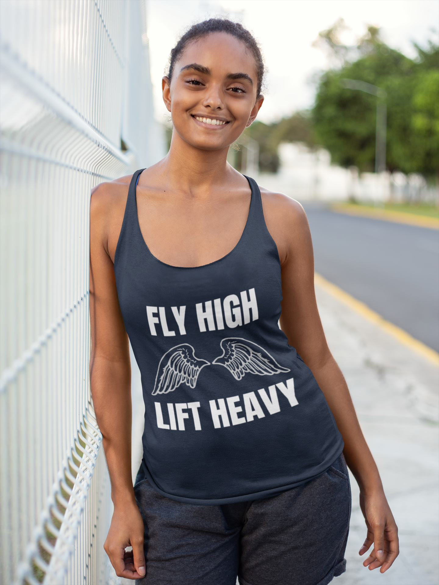 Woman wearing a black tank top with 'Fly High Lift Heavy' text and wings graphic, standing outdoors.